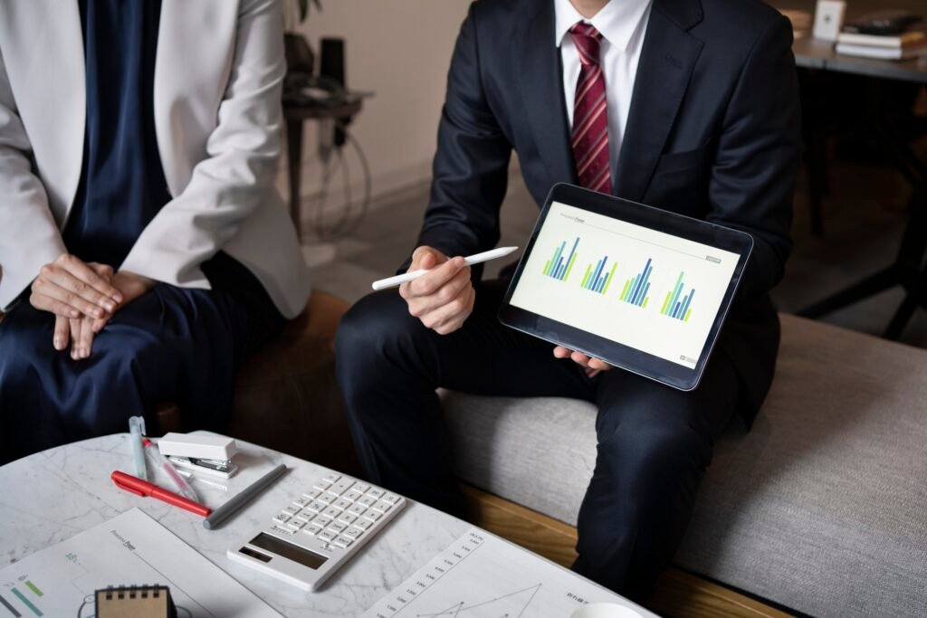 Two business professionals in formal attire discussing data analysis. One person in a suit and red tie holds a tablet displaying bar charts, while the other, dressed in a white blazer, listens. A table with office supplies, a calculator, and printed charts is in front of them.