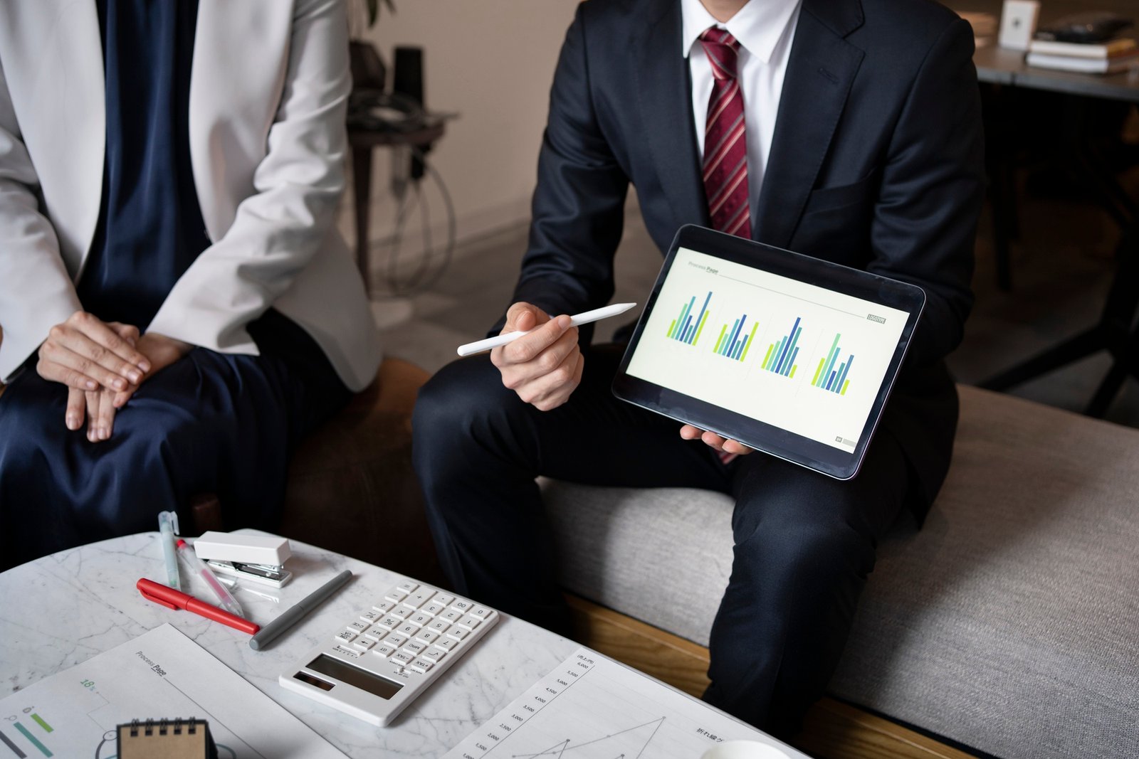 Two business professionals in formal attire discussing data analysis. One person in a suit and red tie holds a tablet displaying bar charts, while the other, dressed in a white blazer, listens. A table with office supplies, a calculator, and printed charts is in front of them.